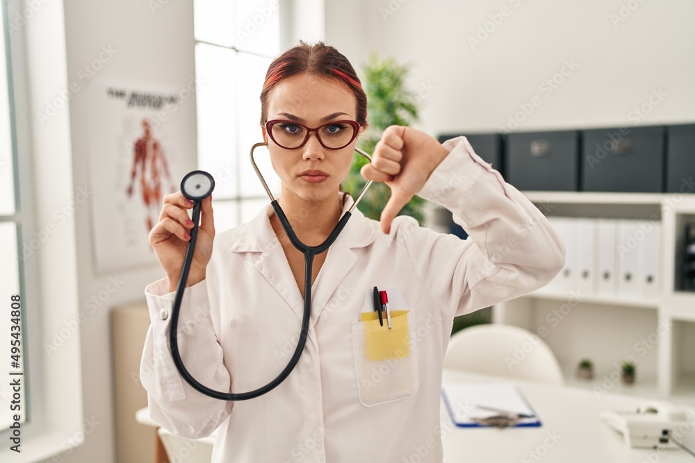 Young caucasian woman wearing doctor uniform using stethoscope with angry face, negative sign showing dislike with thumbs down, rejection concept Young caucasian woman wearing doctor uniform using stethoscope with angry face, negative sign showing dislike with thumbs down, rejection concept
