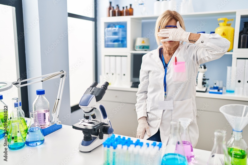Middle age blonde woman working at scientist laboratory covering eyes with hand, looking serious and sad. sightless, hiding and rejection concept Middle age blonde woman working at scientist laboratory covering eyes with hand, looking serious and sad. sightless, hiding and rejection concept