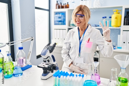 Middle age blonde woman working at scientist laboratory showing and pointing up with fingers number three while smiling confident and happy.