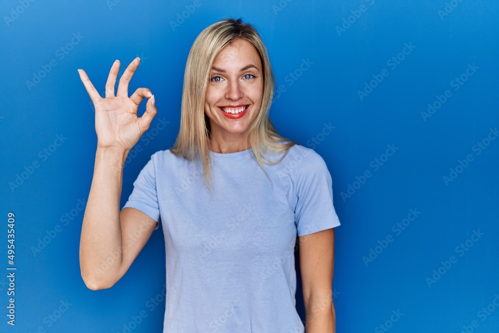 Beautiful blonde woman wearing casual t shirt over blue background smiling positive doing ok sign with hand and fingers. successful expression. Beautiful blonde woman wearing casual t shirt over blue background smiling positive doing ok sign with hand and fingers. successful expression.