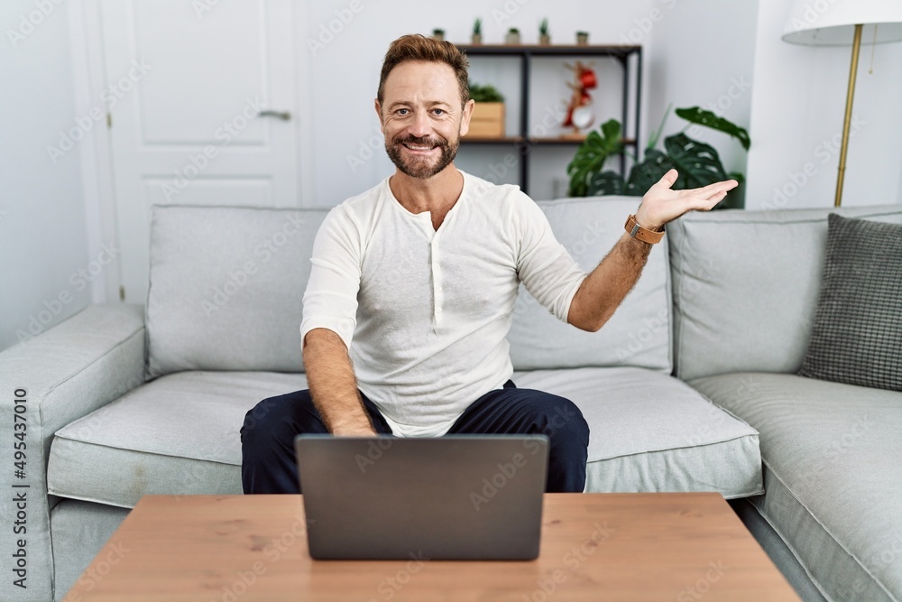 Middle age man using laptop at home smiling cheerful presenting and pointing with palm of hand looking at the camera. Middle age man using laptop at home smiling cheerful presenting and pointing with palm of hand looking at the camera.