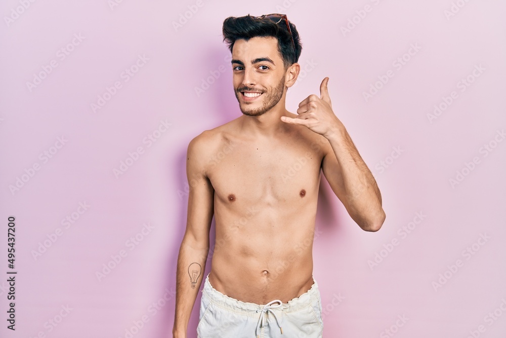 Young hispanic man wearing swimwear shirtless smiling doing phone gesture with hand and fingers like talking on the telephone. communicating concepts.
