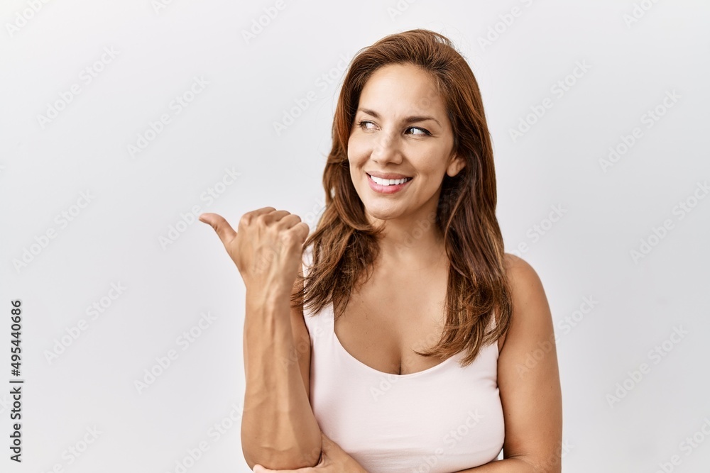 Middle age hispanic woman standing over isolated background smiling with happy face looking and pointing to the side with thumb up. Middle age hispanic woman standing over isolated background smiling with happy face looking and pointing to the side with thumb up.