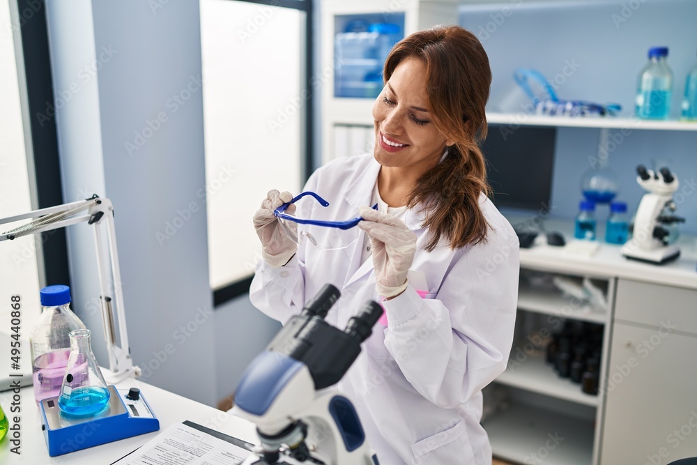 Young latin woman wearing scientist uniform holding safety glasses at laboratory Young latin woman wearing scientist uniform holding safety glasses at laboratory
