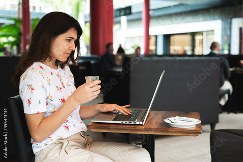 Side view casual young woman sitting at cafe table using computer laptop while drinking a cup of coffee.