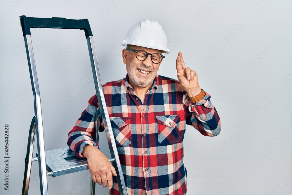 Handsome mature handyman close to construction stairs wearing hardhat gesturing finger crossed smiling with hope and eyes closed. luck and superstitious concept. Handsome mature handyman close to construction stairs wearing hardhat gesturing finger crossed smiling with hope and eyes closed. luck and superstitious concept.