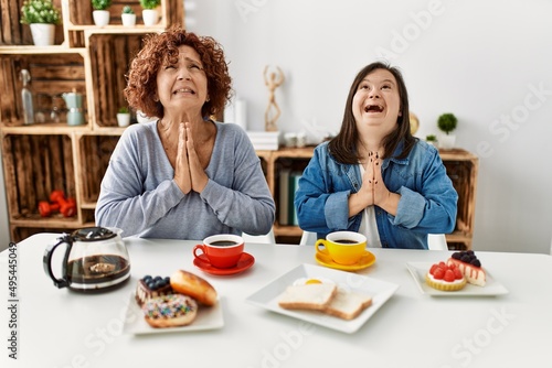 Family of mother and down syndrome daughter sitting at home eating breakfast begging and praying with hands together with hope expression on face very emotional and worried. begging.