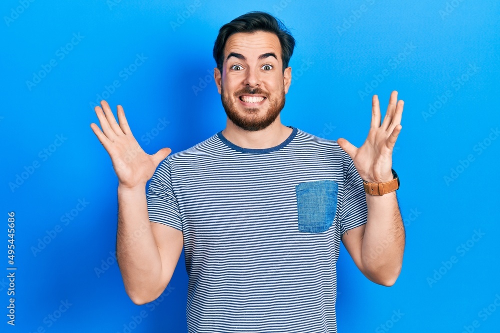 Handsome caucasian man with beard wearing casual striped t shirt celebrating crazy and amazed for success with arms raised and open eyes screaming excited. winner concept Handsome caucasian man with beard wearing casual striped t shirt celebrating crazy and amazed for success with arms raised and open eyes screaming excited. winner concept