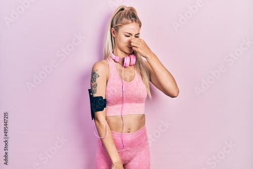 Young beautiful caucasian woman wearing gym clothes and using headphones smelling something stinky and disgusting, intolerable smell, holding breath with fingers on nose. bad smell