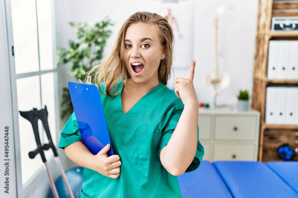 Young caucasian woman working at pain recovery clinic pointing finger up with successful idea. exited and happy. number one. Young caucasian woman working at pain recovery clinic pointing finger up with successful idea. exited and happy. number one.