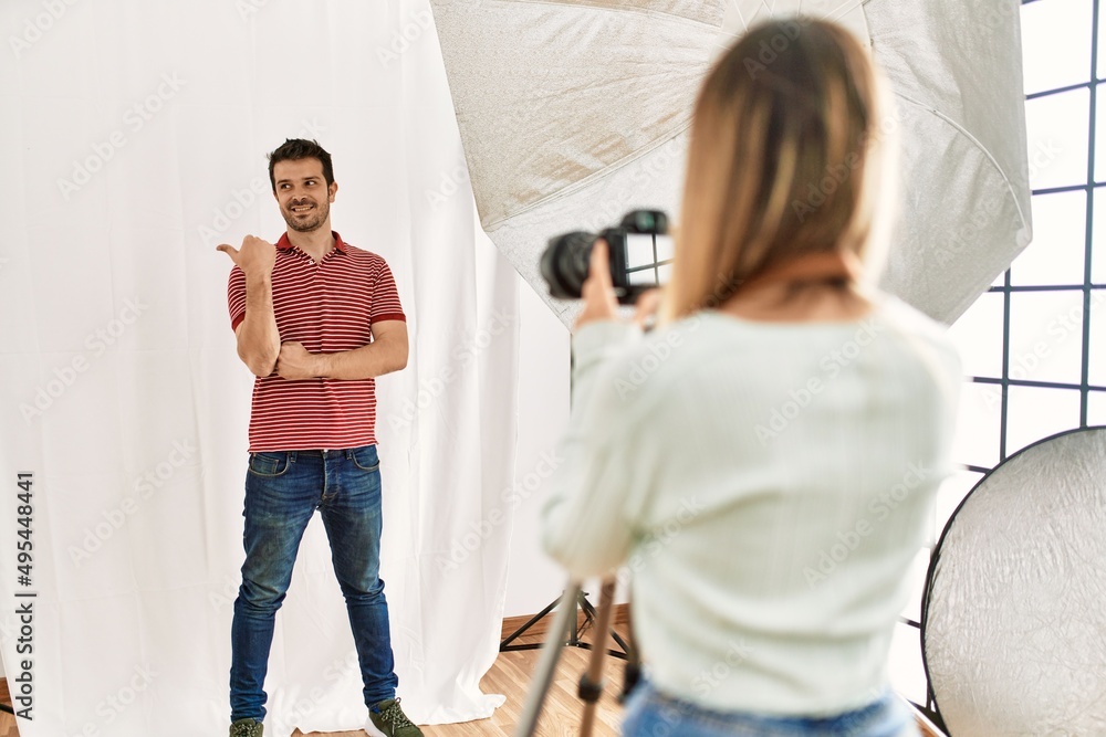 Woman photographer talking pictures of man posing as model at photography studio smiling with happy face looking and pointing to the side with thumb up. Woman photographer talking pictures of man posing as model at photography studio smiling with happy face looking and pointing to the side with thumb up.