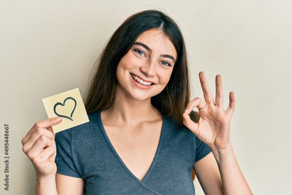 Young caucasian woman holding heart reminder doing ok sign with fingers, smiling friendly gesturing excellent symbol Young caucasian woman holding heart reminder doing ok sign with fingers, smiling friendly gesturing excellent symbol