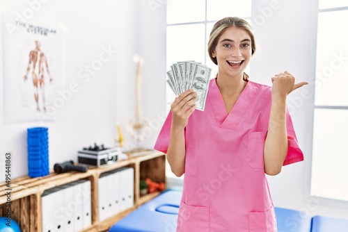 Young physiotherapist woman working at pain recovery clinic holding dollars pointing thumb up to the side smiling happy with open mouth
