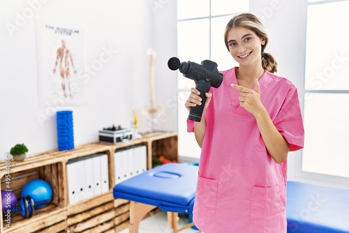 Young physiotherapist woman holding therapy massage gun at wellness center smiling happy pointing with hand and finger