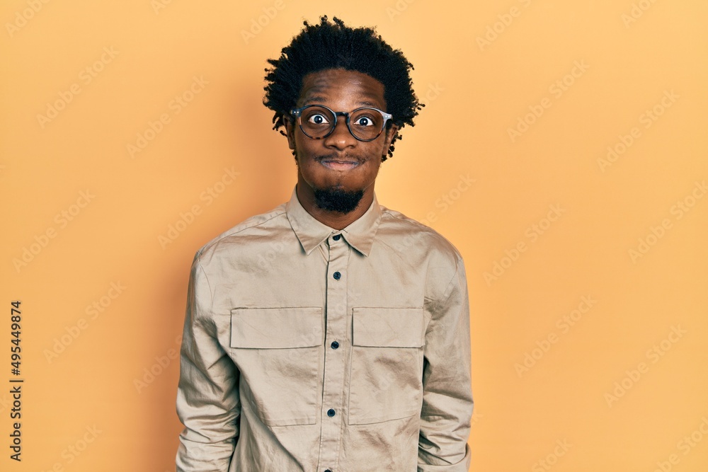 Obraz premium Young african american man wearing casual clothes and glasses puffing cheeks with funny face. mouth inflated with air, crazy expression.