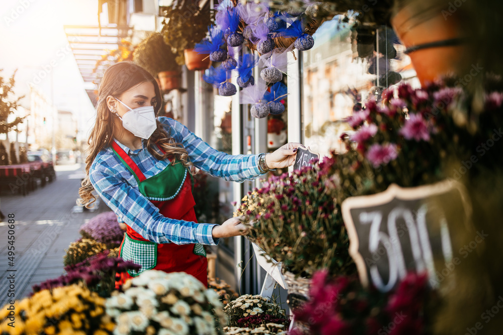 Young adult woman working in city street flower shop or florist. She is wearing protective face mask during Covid-19 virus pandemic. Small business concept.
