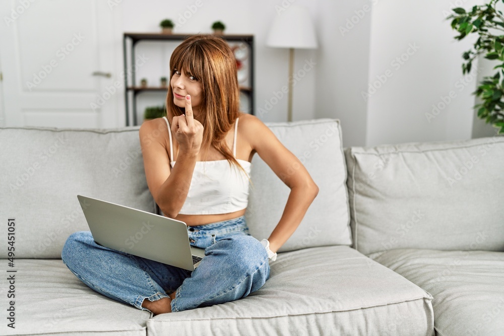 Hispanic woman sitting on the sofa at home using laptop showing middle finger, impolite and rude fuck off expression Hispanic woman sitting on the sofa at home using laptop showing middle finger, impolite and rude fuck off expression