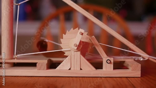 SLOW MOTION: Close up of a toy wooden trebuchet firing on a wooden table with a chair and counter in the background. The firing arm swings away and the counterweight swings into view. photo