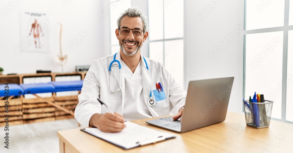 Middle age hispanic therapist man working at pain recovery clinic with laptop looking positive and happy standing and smiling with a confident smile showing teeth