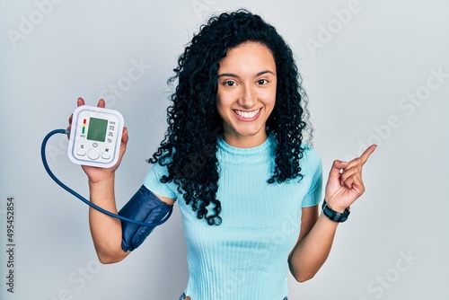 Young hispanic woman with curly hair using blood pressure monitor smiling happy pointing with hand and finger to the side photo
