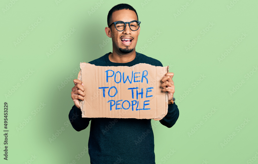 Young african american man holding power to the people banner winking looking at the camera with sexy expression, cheerful and happy face. Young african american man holding power to the people banner winking looking at the camera with sexy expression, cheerful and happy face.