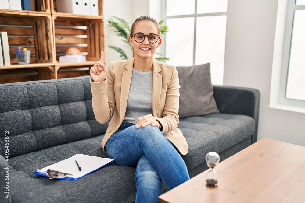 Young woman working at consultation office showing and pointing up with finger number one while smiling confident and happy. Young woman working at consultation office showing and pointing up with finger number one while smiling confident and happy.