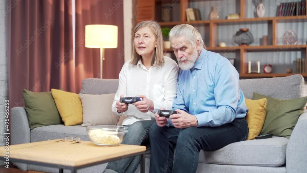 senior couple playing video game console with joysticks sitting on couch in living room. Funny Mature people family having fun spend leisure time together. gray hair man and woman enjoy entertaining senior couple playing video game console with joysticks sitting on couch in living room. Funny Mature people family having fun spend leisure time together. gray hair man and woman enjoy entertaining