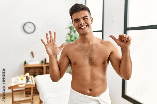 Young hispanic man standing shirtless at spa center showing and pointing up with fingers number six while smiling confident and happy.