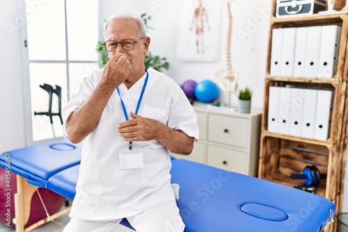 Senior physiotherapy man working at pain recovery clinic smelling something stinky and disgusting  intolerable smell  holding breath with fingers on nose. bad smell