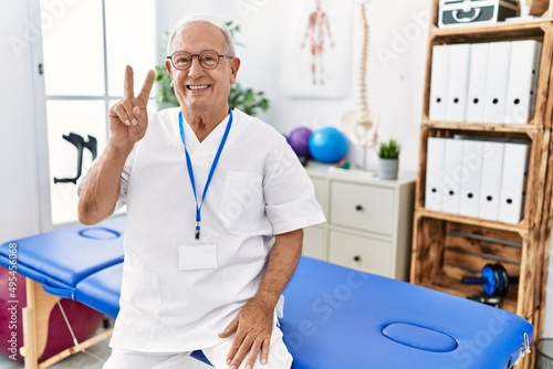 Senior physiotherapy man working at pain recovery clinic showing and pointing up with fingers number two while smiling confident and happy.