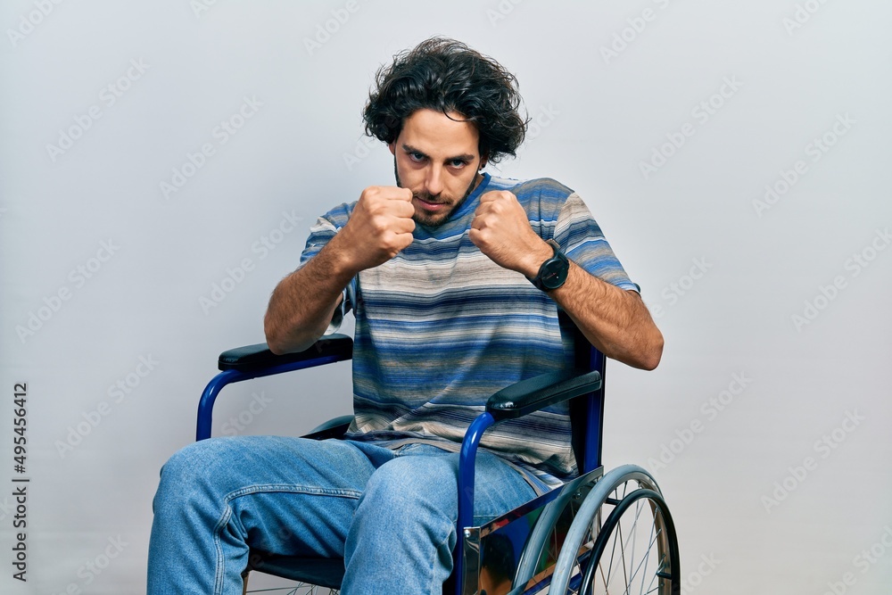 Handsome hispanic man sitting on wheelchair ready to fight with fist defense gesture, angry and upset face, afraid of problem Handsome hispanic man sitting on wheelchair ready to fight with fist defense gesture, angry and upset face, afraid of problem
