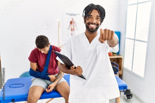 Young hispanic man working at pain recovery clinic with a man with broken arm pointing to you and the camera with fingers  smiling positive and cheerful