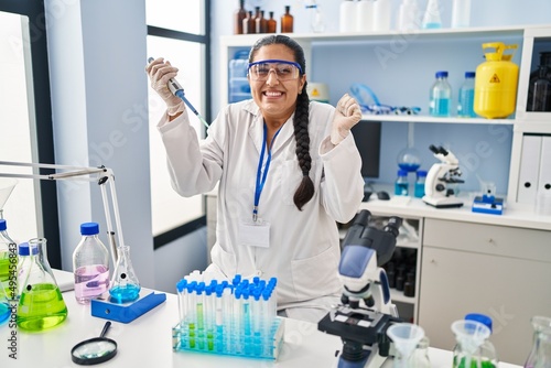 Young hispanic woman working at scientist laboratory screaming proud  celebrating victory and success very excited with raised arm