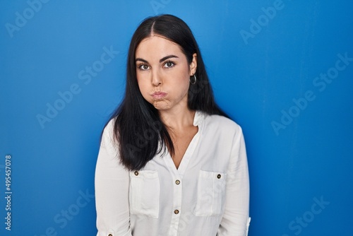 Young hispanic woman standing over blue background puffing cheeks with funny face. mouth inflated with air, crazy expression.