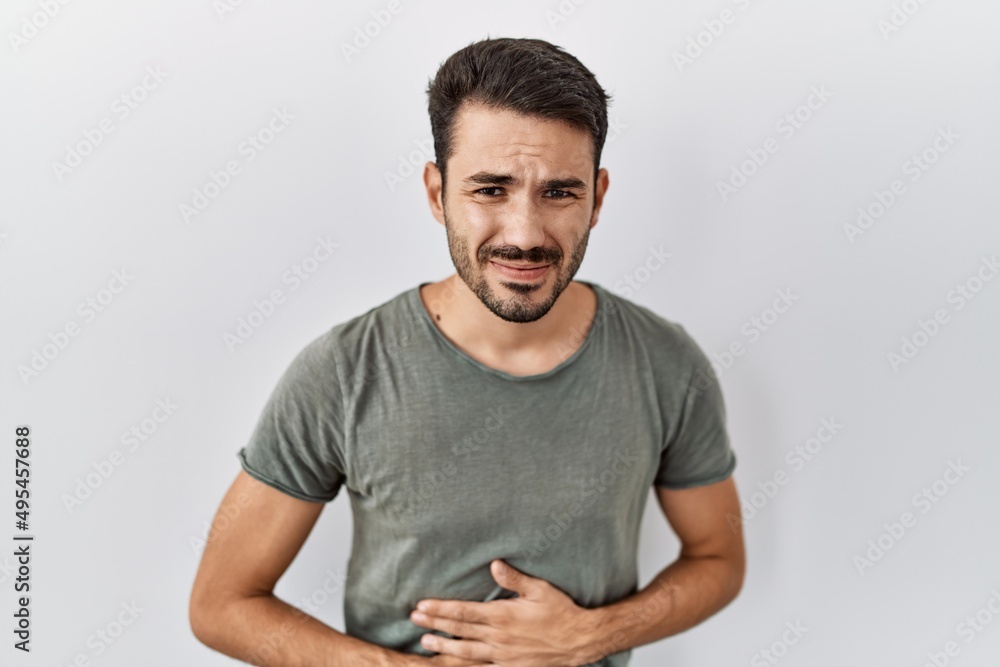Young hispanic man with beard wearing casual t shirt over white background with hand on stomach because indigestion, painful illness feeling unwell. ache concept. Young hispanic man with beard wearing casual t shirt over white background with hand on stomach because indigestion, painful illness feeling unwell. ache concept.