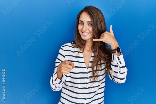 Young hispanic woman standing over blue isolated background smiling doing talking on the telephone gesture and pointing to you. call me. © Krakenimages.com