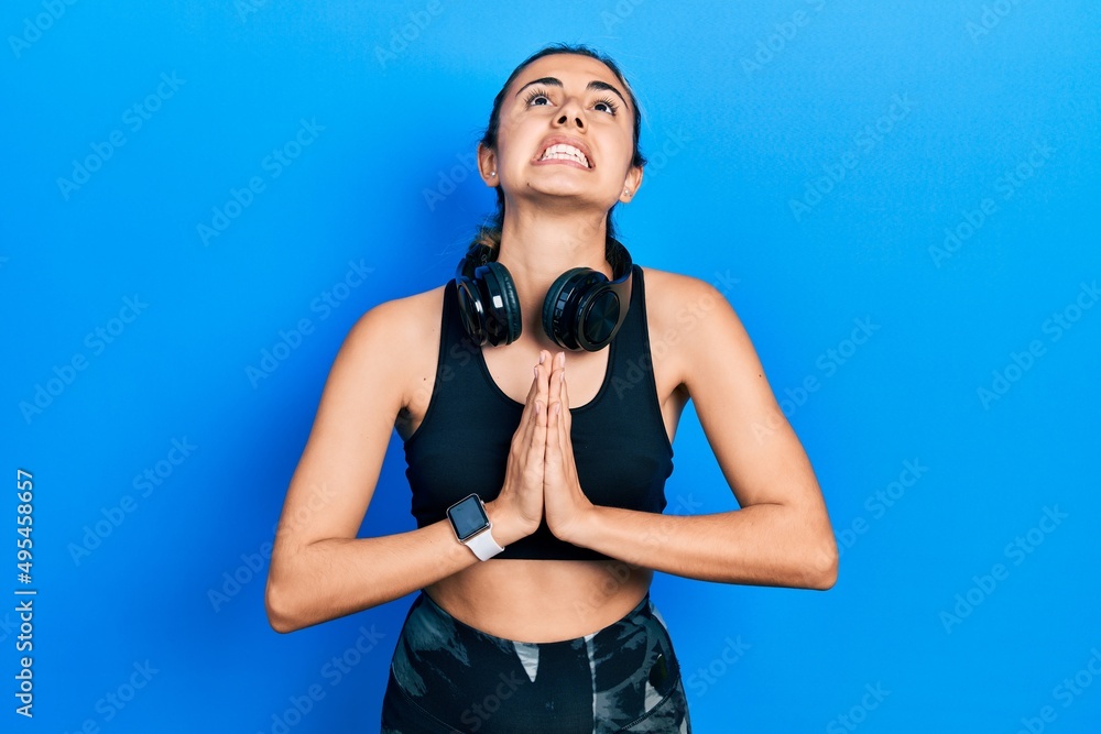 Obraz premium Beautiful hispanic woman wearing gym clothes and using headphones begging and praying with hands together with hope expression on face very emotional and worried. begging.