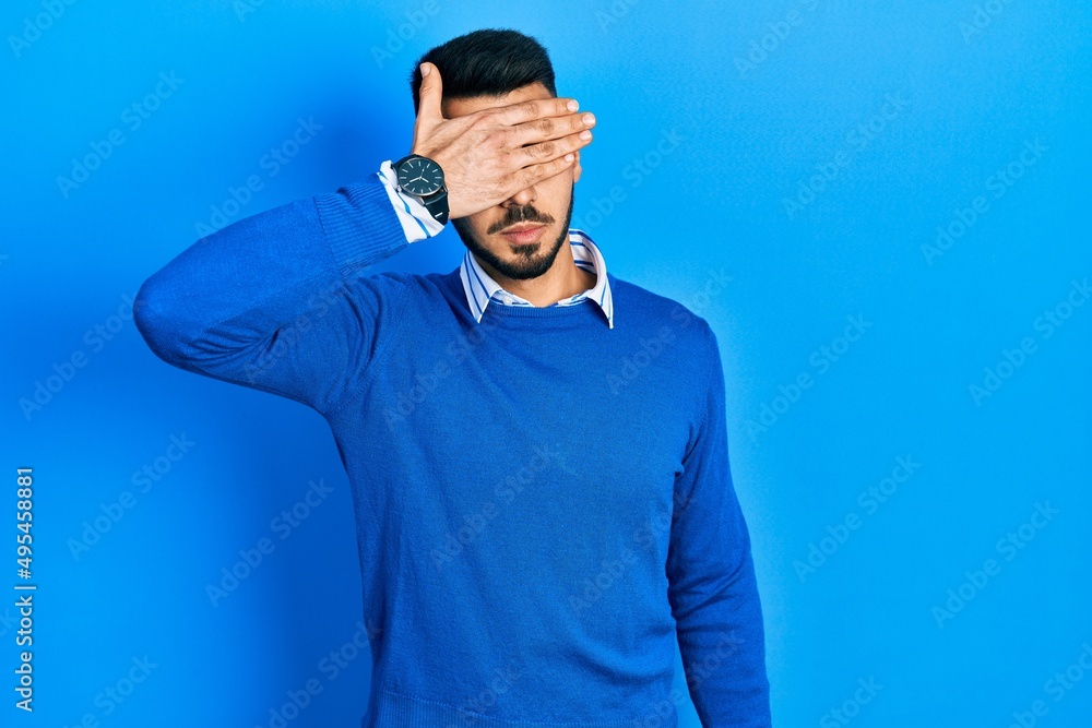Fototapeta premium Young hispanic man with beard wearing casual blue sweater covering eyes with hand, looking serious and sad. sightless, hiding and rejection concept