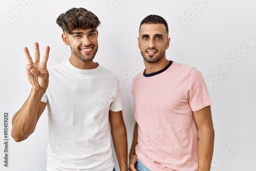 Young gay couple standing over isolated background showing and pointing up with fingers number three while smiling confident and happy.