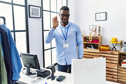 Young african man working as manager at retail boutique waiving saying hello happy and smiling, friendly welcome gesture