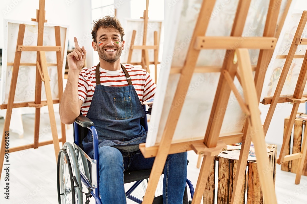 Young handsome man with beard at art studio sitting on wheelchair surprised with an idea or question pointing finger with happy face, number one