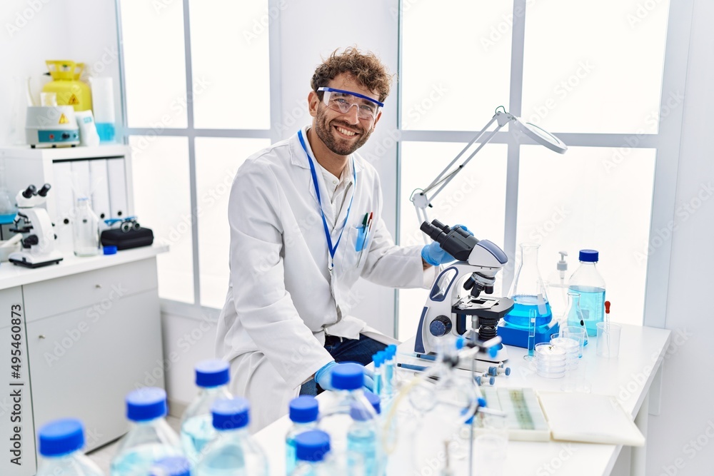 Fototapeta premium Young hispanic man wearing scientist uniform using microscope at laboratory