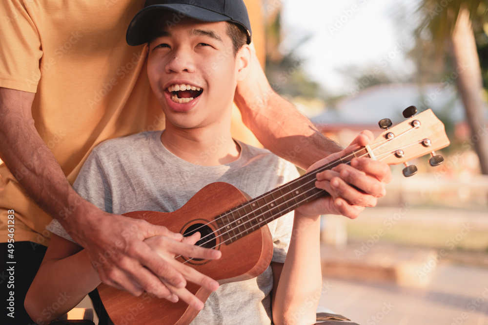 Obraz premium Happy face of young man with disability holding ukulele and singing, playing with music therapy on the outdoor nature background,Vacation hobby activity with family activity and mental health concept.