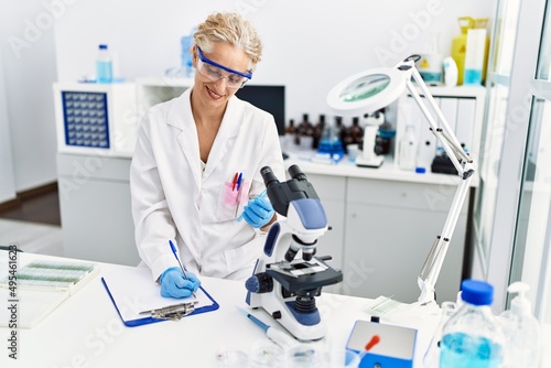 Middle age blonde woman wearing scientist uniform holding test tube writing on clipboard at laboratory