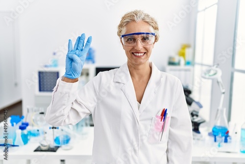 Middle age blonde woman working at scientist laboratory showing and pointing up with fingers number four while smiling confident and happy.
