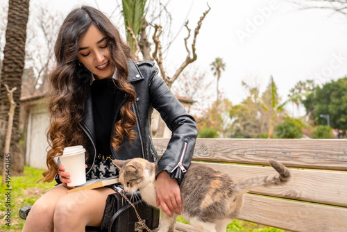 Animal love  young woman sitting in the park loves the stray cat that comes to her  she is holding a cardboard coffee cup and loves a cat  a public park and street animal