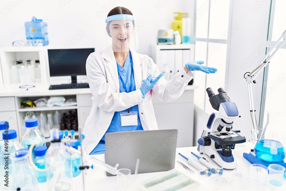 Young hispanic woman wearing scientist uniform amazed and smiling to the camera while presenting with hand and pointing with finger. Young hispanic woman wearing scientist uniform amazed and smiling to the camera while presenting with hand and pointing with finger.