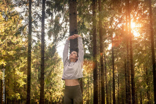 Woman stretching arms and breathing fresh air in middle of a forest while exercising at sunrise.