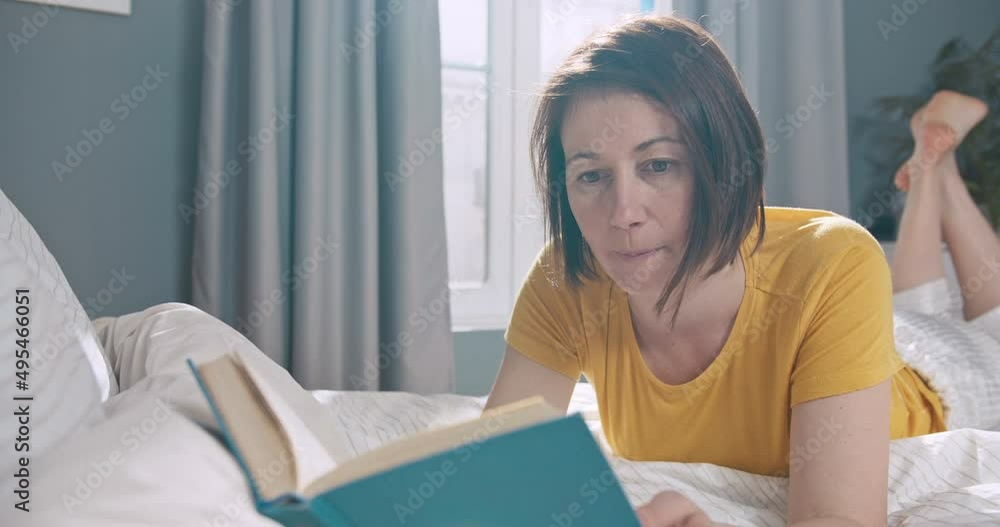 Attractive caucasian female lying on the bed and reading a book in her apartment. Young woman enjoying her time lying on the bed in the morning. Attractive caucasian female lying on the bed and reading a book in her apartment. Young woman enjoying her time lying on the bed in the morning.