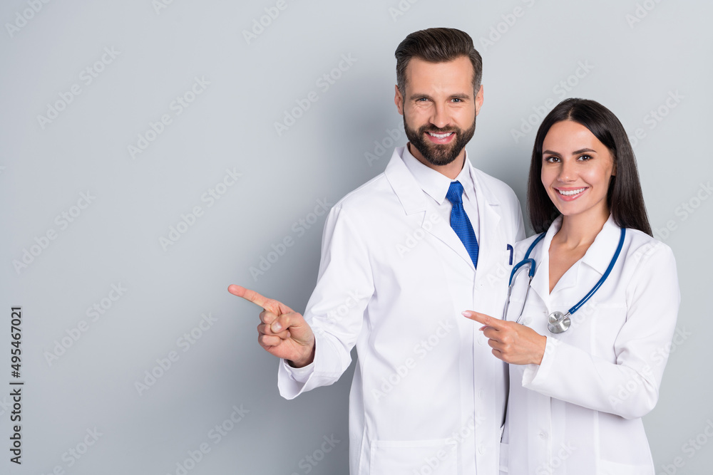 Photo of positive pretty two doctors wear white coats pointing empty space hugging isolated grey color background Photo of positive pretty two doctors wear white coats pointing empty space hugging isolated grey color background